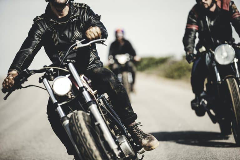 Three men wearing leather jackets riding cafe racer motorcycles along rural road.